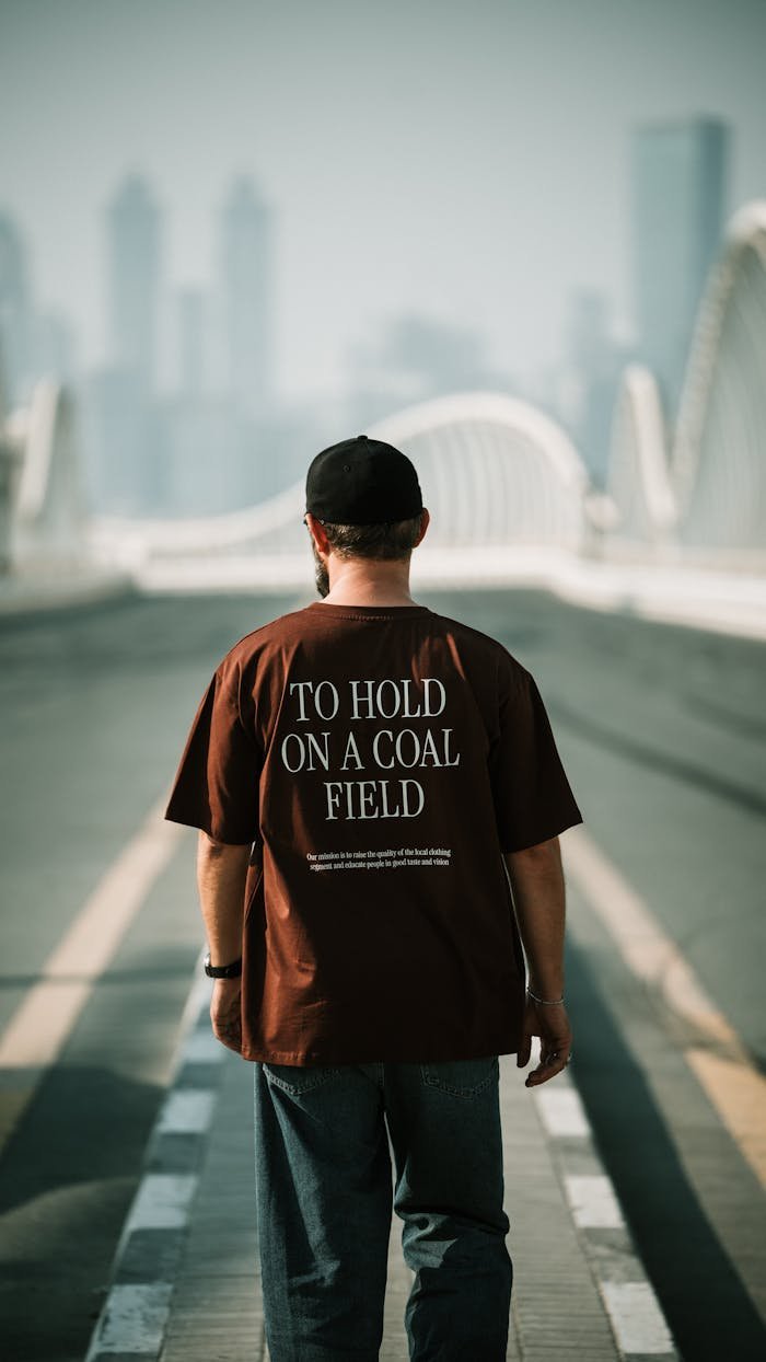 A man in a graphic tee walks on an urban bridge with a city skyline in the background.